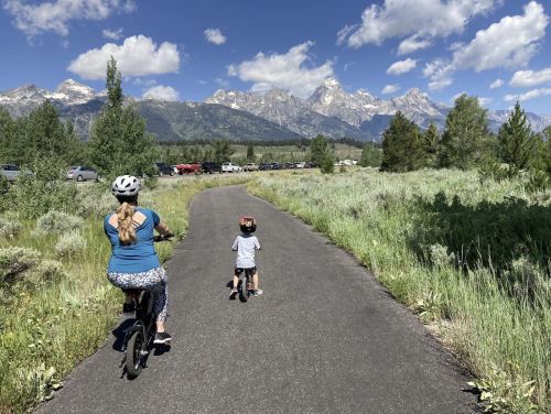 A woman and a child ride bikes on a path with mountains in the background and greenery on either side.