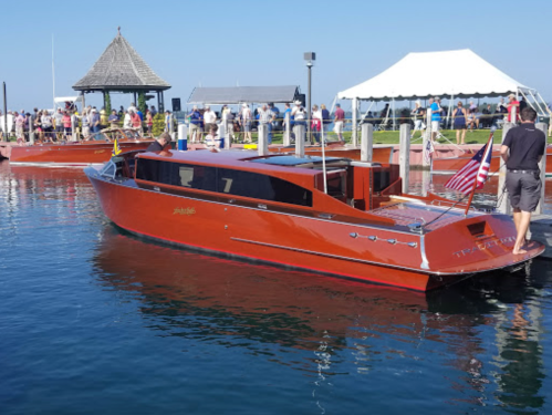 A vintage wooden boat docked at a marina with people in the background and a tent set up nearby.