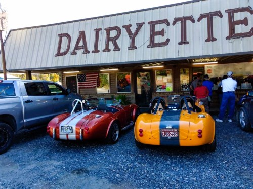 Two classic sports cars parked outside a diner named "Dairyette," with people inside ordering food.