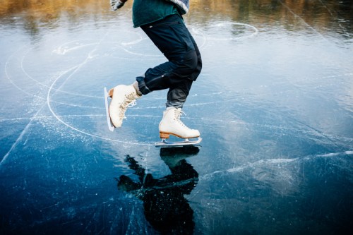 A person skating on a frozen lake, wearing ice skates and black pants, with a reflection on the ice surface.