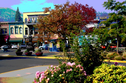Colorful buildings line a street, framed by trees and blooming flowers, with parked cars in the background.