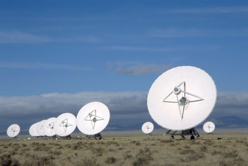 A row of large satellite dishes facing the sky in a desert landscape under a cloudy blue sky.