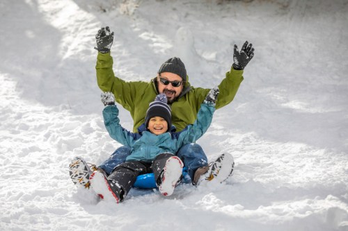 A man and a boy joyfully sledding down a snowy hill, both smiling and raising their arms in excitement.