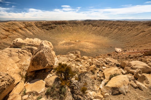 A vast, barren crater surrounded by rocky terrain under a clear blue sky, with small figures visible in the distance.