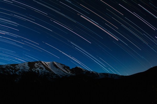 Star trails streak across a night sky above snow-capped mountains, creating a stunning celestial display.