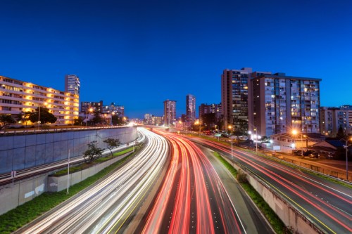 City skyline at dusk with light trails from moving traffic on a highway and illuminated buildings in the background.
