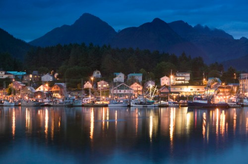 A serene harbor at dusk, with boats and buildings reflecting on calm water, surrounded by mountains.