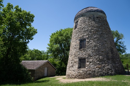 A tall, stone windmill stands beside a small, rustic building, surrounded by lush green trees under a clear blue sky.