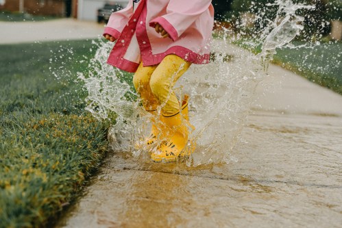 A child in a pink raincoat and yellow boots splashes in a puddle on a rainy day.