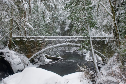 A snow-covered stone bridge arches over a river, surrounded by trees blanketed in fresh snow.