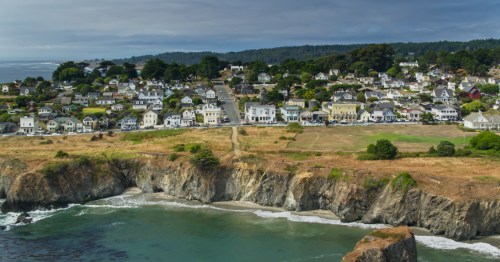 A coastal town with white houses on a hillside, overlooking a rocky shore and calm ocean under a cloudy sky.
