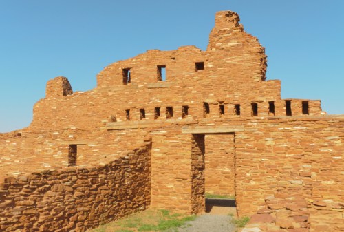 Ancient stone ruins with multiple windows against a clear blue sky, surrounded by grassy areas.
