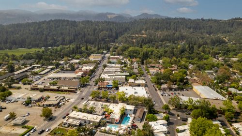 Aerial view of a small town surrounded by green hills, featuring shops, parking lots, and residential areas.