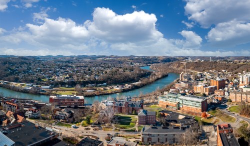 A panoramic view of a river winding through a valley, surrounded by hills and buildings under a blue sky with clouds.