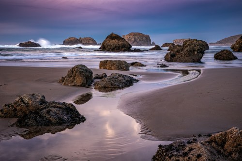 A serene beach scene with rocks, gentle waves, and a winding stream reflecting the sky at dusk.