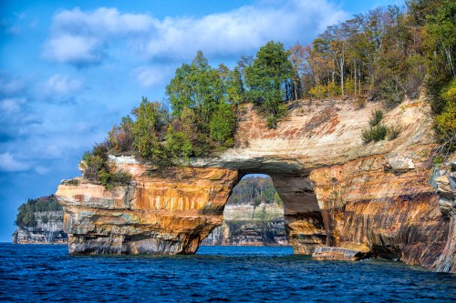A natural rock arch stands over blue water, surrounded by colorful trees and cliffs under a partly cloudy sky.