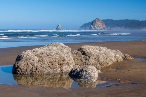 A sandy beach with large rocks, gentle waves, and distant cliffs under a clear blue sky.