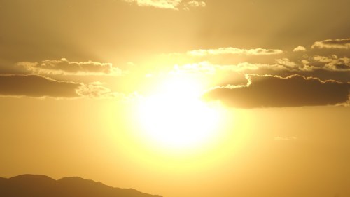 A bright sun setting behind clouds, casting a warm golden glow over distant mountains.