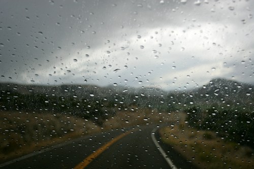 Raindrops on a car windshield with a winding road and cloudy mountains in the background.