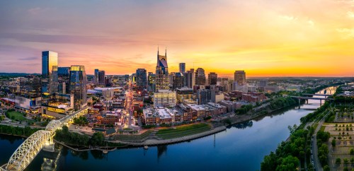 A panoramic view of Nashville's skyline at sunset, with the river and city lights reflecting in the water.