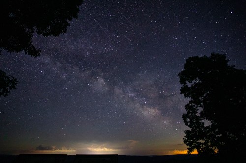 A starry night sky filled with the Milky Way, framed by silhouetted trees and distant glowing clouds.