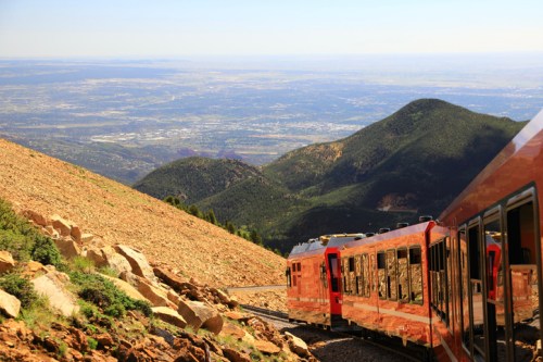 A red train winds along a rocky mountainside, overlooking a vast valley and distant mountains under a clear blue sky.