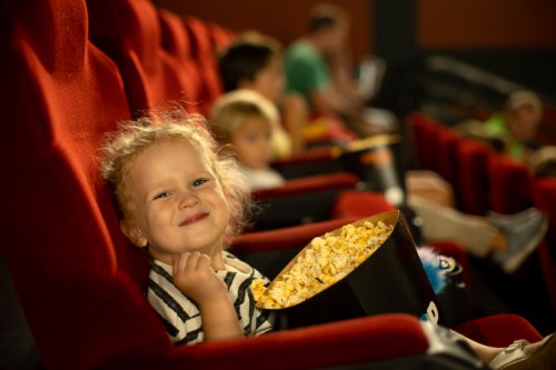 A smiling child holds a bucket of popcorn in a movie theater, with other kids seated in the background.