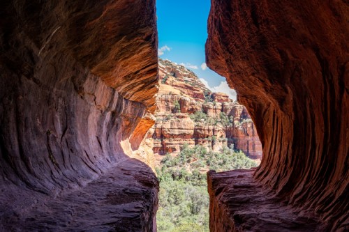 A scenic view through a rock formation, showcasing red cliffs and lush greenery under a bright blue sky.