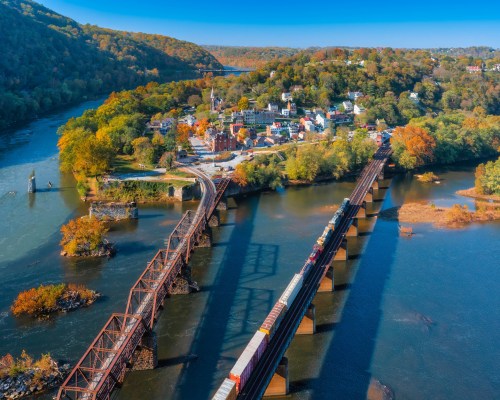 Aerial view of a scenic river with train tracks, colorful autumn trees, and a small town nestled in the landscape.