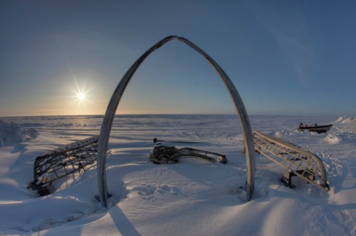A whale bone arch stands in a snowy landscape, with the sun setting over a frozen sea in the background.
