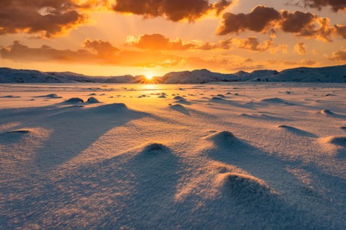 A serene winter landscape with snow-covered mounds and a vibrant sunset illuminating the horizon.