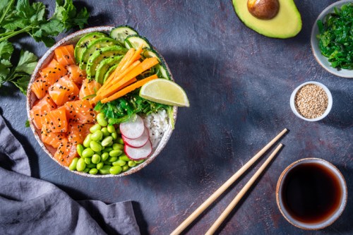 A colorful poke bowl with salmon, avocado, edamame, carrots, and rice, garnished with lime and sesame seeds.