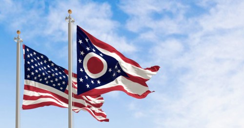 Two flags waving in the wind: the American flag and the Ohio state flag against a blue sky.