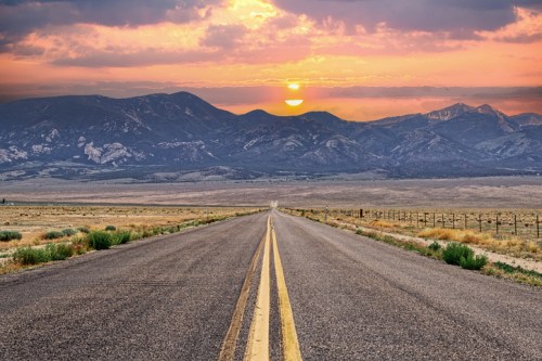 A winding road stretches toward a sunset behind mountains, with a vast landscape and cloudy sky.