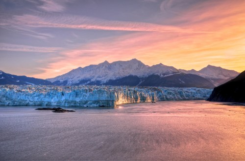 A stunning sunset over a glacier, reflecting vibrant colors on the water, with mountains in the background.