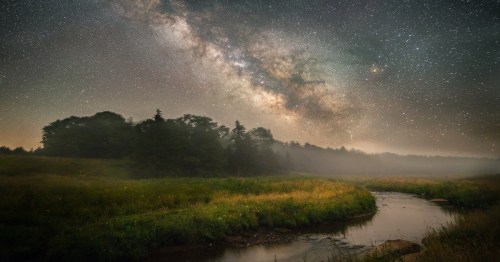 A serene landscape featuring a river, lush grass, and a starry night sky with the Milky Way prominently displayed.
