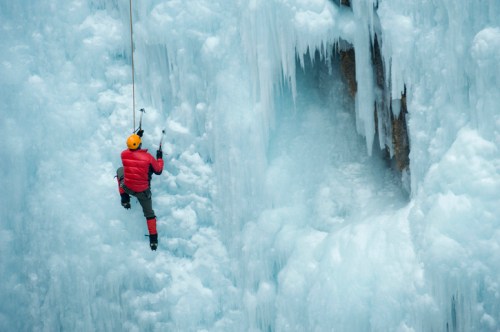 A climber in a red jacket ascends a frozen waterfall, surrounded by icy formations and a rocky cliff.