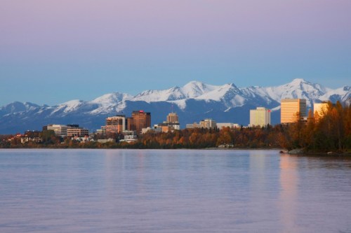 City skyline with modern buildings against a backdrop of snow-capped mountains and a calm lake at sunset.