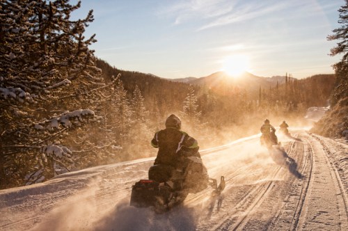 Two snowmobiles ride down a snowy trail at sunset, surrounded by trees and mountains. Dust rises in the golden light.
