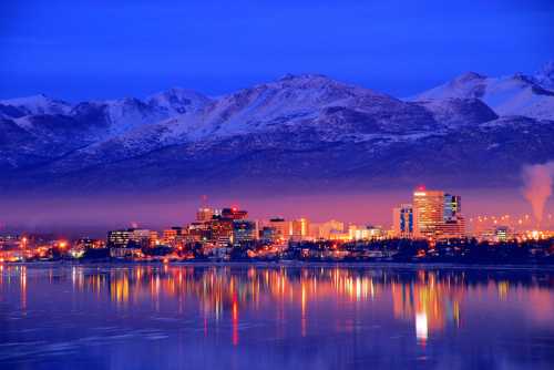 A city skyline at dusk, reflecting on water, with snow-capped mountains in the background and lights twinkling.