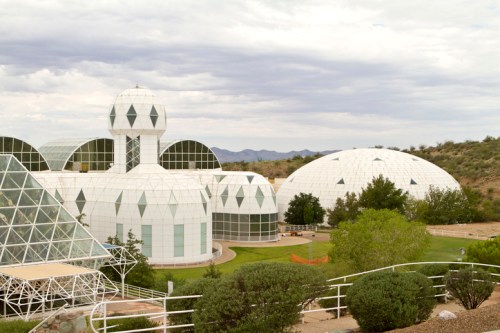 Geodesic dome structures surrounded by greenery under a cloudy sky in a desert landscape.