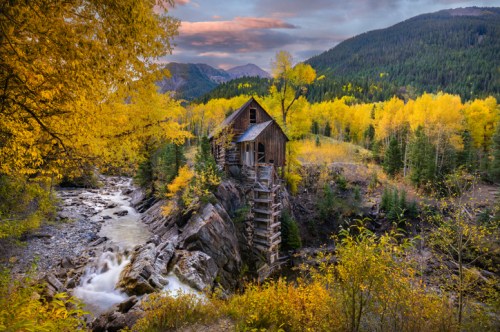 A rustic wooden cabin surrounded by vibrant autumn foliage and a flowing river, set against a mountainous backdrop.
