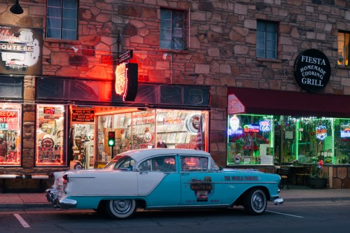 A vintage turquoise and white car parked in front of a retro diner with neon signs and a stone building backdrop.