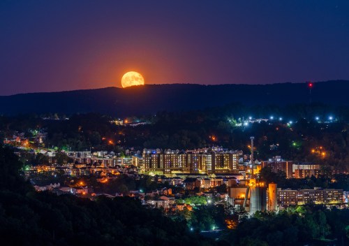 A vibrant cityscape at night with a large full moon rising over the hills in the background.