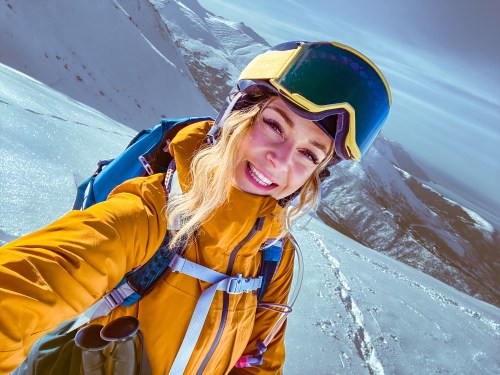 A smiling woman in a yellow jacket and helmet takes a selfie on a snowy mountain slope.