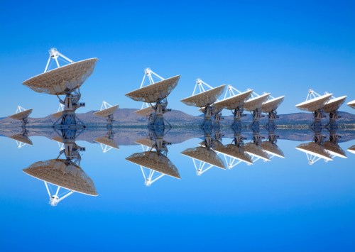 A row of large satellite dishes against a clear blue sky, reflecting on calm water below.