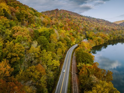 A winding road alongside a lake, surrounded by vibrant autumn foliage and rolling hills.