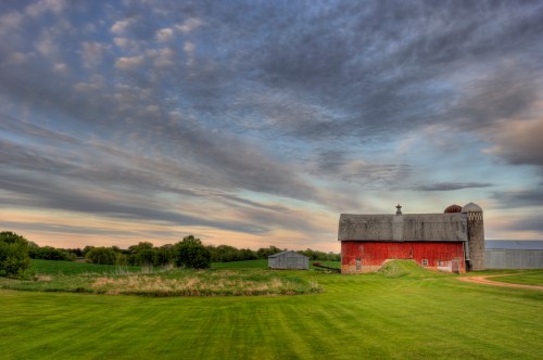 A red barn stands in a green field under a dramatic sky with clouds at sunset.
