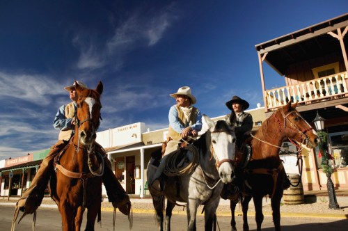 Three cowboys on horseback in a western town, with buildings and a blue sky in the background.