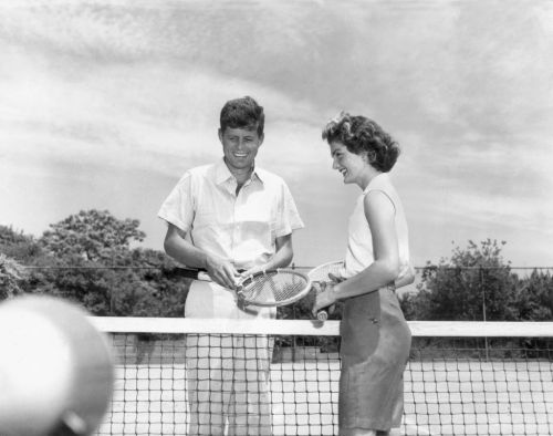 A young man and woman smile at each other while holding tennis rackets on a court.